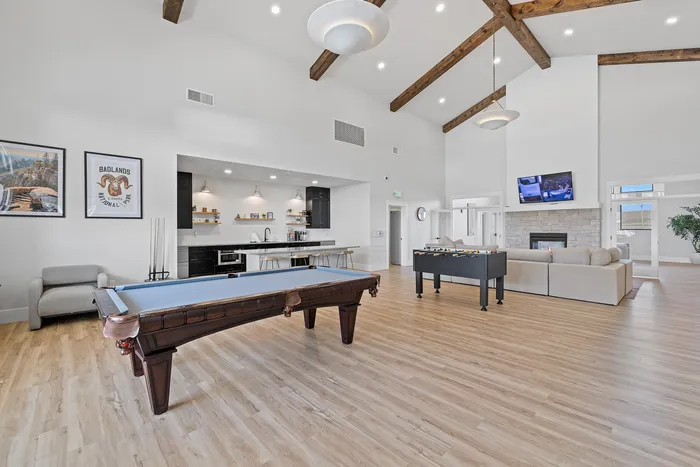 Spacious recreational room with high ceilings, featuring a stylish pool table, cozy sitting area with fireplace, and a modern kitchen space.