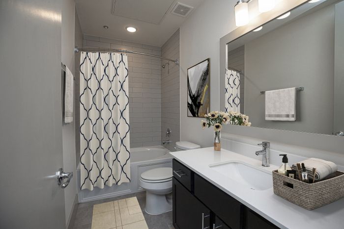 Elegant modern bathroom featuring sleek black cabinetry, a spacious white countertop with stylish accessories, decorative floral arrangement, and a contemporary shower area with patterned curtain and ambient lighting.