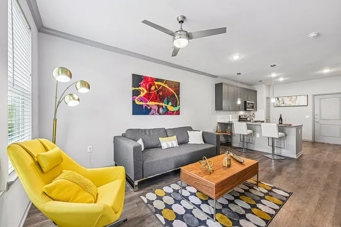 Modern living room with vibrant decor, featuring a cozy gray sofa, bright yellow armchair, stylish artwork, and a sleek open kitchen with a breakfast bar.