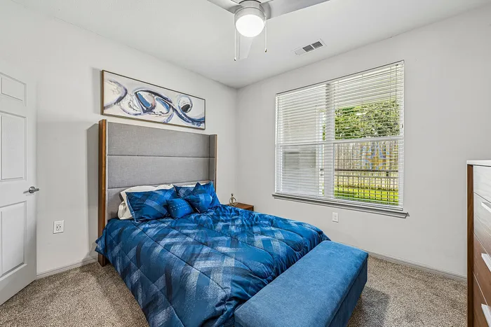 Chic bedroom featuring a plush bed with blue linens, modern art, and ample natural light from large windows.