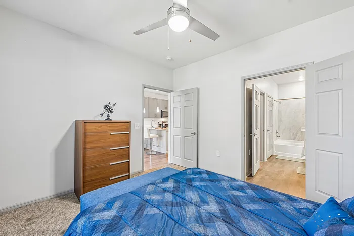 Bright bedroom featuring modern ceiling fan, cozy blue bedding, sleek wooden dresser, with a view into a stylish bathroom and elegant kitchen area.