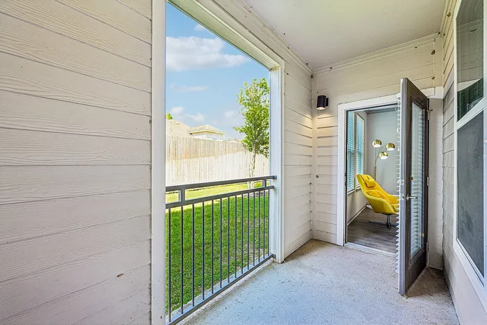 Charming apartment balcony with a sleek metal railing, offering a view of a neatly maintained garden and a glimpse into a stylish interior featuring a vibrant yellow chair and modern lighting.