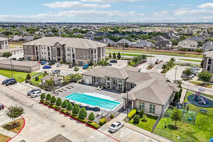 Aerial view of a modern apartment complex featuring a sparkling blue swimming pool, lush landscaping, and a children's playground, set in a peaceful suburban neighborhood.