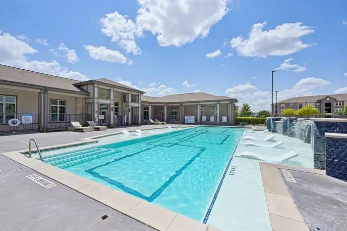 Luxurious apartment community pool with elegant lounge chairs and waterfall feature under a bright blue sky.