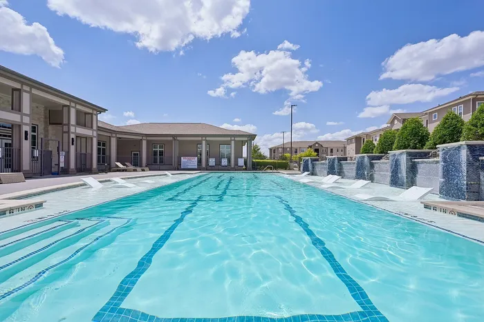 Luxurious apartment complex showcasing a pristine swimming pool with waterfall features and modern poolside seating under a bright blue sky.