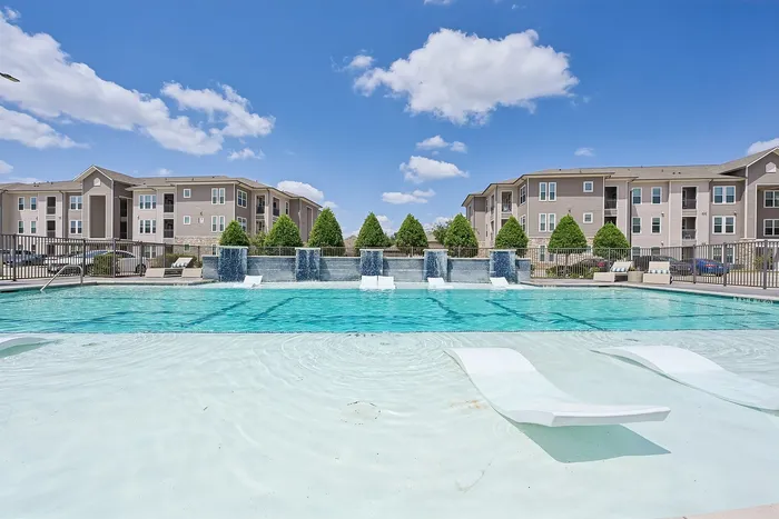 Luxurious apartment complex featuring a stunning pool with sun loungers and tranquil water features, set against modern buildings and a vibrant blue sky.