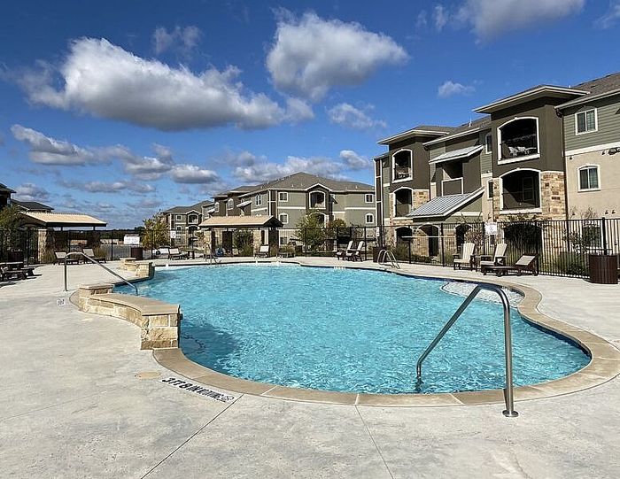 Inviting luxury apartment complex with a sparkling blue swimming pool surrounded by modern beige and stone-accented buildings under a bright, clear sky.