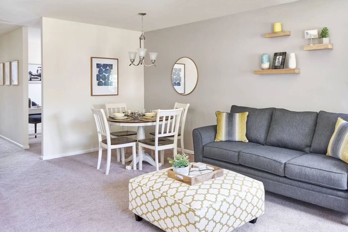 Pet-Friendly Apartments in Bensalem, PA - Village Square - Living Room with Carpeted Flooring, Grey Couch, Cushioned Coffee Table, Floating Display Shelves, Dining Nook with Table and Four Chairs, Round Wall Mirror, and Silver Chandelier.