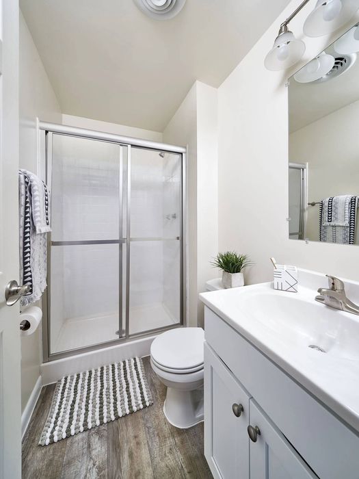 Dog-Friendly Apartments in Millersville, PA - Millers Crossing - White Bathroom with Wood-Style Flooring, Walk-In Shower with Sliding Glass Door, Silver Towel Rack, White Vanity with Sink and Cabinetry, and Large Mirror.