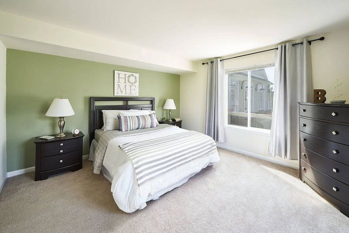 Millersville, PA, Apartments Near Millersville University - Millers Crossing - Furnished Bedroom with Plush Carpet Flooring, Large Bed, Wooden Nightstands with Lamps and Drawers, Wooden Dresser, Pale Green Accent Wall, and Large Window.