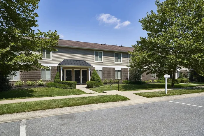 Apartments for Rent in Millersville, PA - Millers Crossing - Street View of Property Exterior with Brick Building with Tan Walls, Windows, White Trim, Black Doors, Concrete Path, and Lush Greenery.