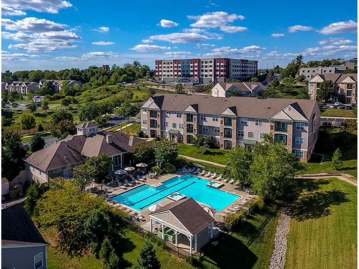 Dog-Friendly Apartments in Plymouth Meeting, PA - Plymouth Pointe - Aerial View of Property Showing Apartment Building, Clubhouse, Pool with Pavilion, Lounge Seats, Umbrella Tables and Chairs, and Lush Greenery.