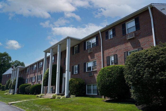 West Chester, PA, Apartments Near West Chester University - Goshen Manor - Brick Property Exterior with White Trim and Columns, Black Window Shutters, Grassy Lawns, and Large Manicured Hedges.