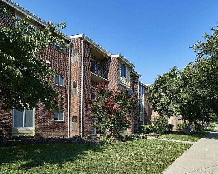 Frederick, MD, Apartments - Willowdale Crossing - Property Exterior with Brick Walls, Manicured Grass Lawn, Flowering Plant, Trees, and Concrete Walking Path.
