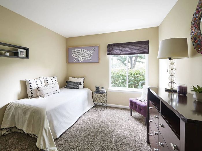 One-Bedroom Apartments in Frederick, MD - Willowdale Crossing - Bedroom with Carpeted Flooring, Bed, Round Nightstand, Brown Dresser with Lamp, Purple Chair, and Window.