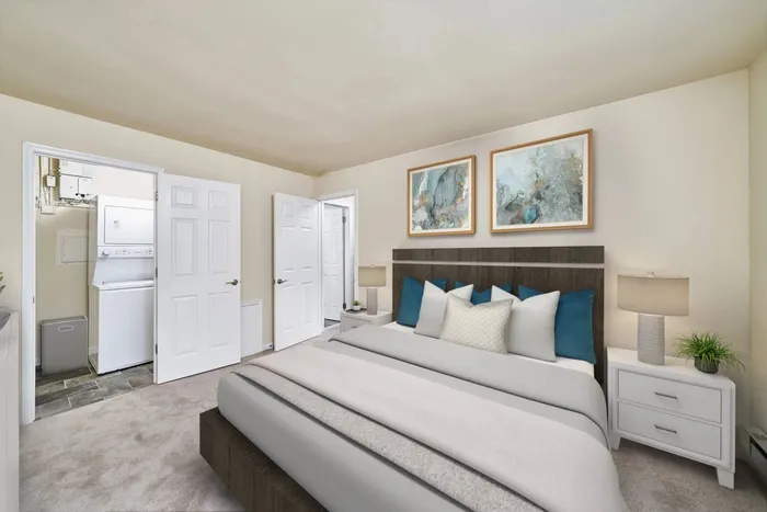 Dublin, PA Apartments - Dublin Village - Furnished Bedroom with Carpeted Flooring, Bed, White Nightstands with Drawers and Lamps, Beige Walls, Mounted Art, and Attached Laundry Room with Tiled Flooring.