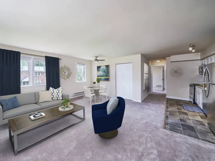 Pet-Friendly Apartments in Dublin, PA - Dublin Village - Spacious Living Room with Carpeted Flooring, Grey Couch, Long Rectangular Coffee Table, Blue Velvet Armchair, Windows, Round Dining Table with Chairs, Ceiling Fan, and View of Tiled Kitchen Area.