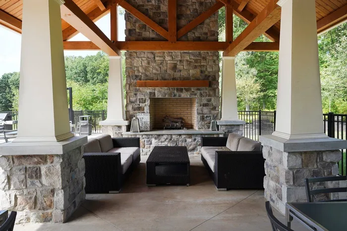 Apartments for Rent State College, PA - Toftrees - Outdoor Picnic Pavilion with Cushioned Furniture, Wood-Buring Fireplace, Stone Details, Vaulted Wooden Ceiling, and View of Nearby Trees.