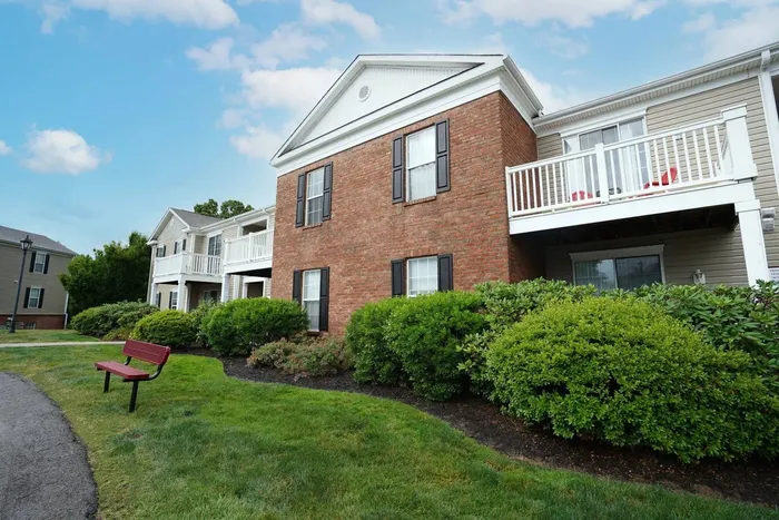 Apartments for Rent in Pittsburgh PA - Bear Run Village - Property Exterior with Brick and Tan Walls, Balconies with White Guardrails, Black Window Shutters, Landscaped Hedges, Grassy Lawns, a Red Bench, and Concrete Path
