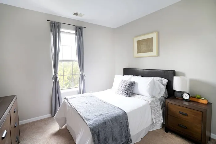 1 BR Apartments in Pittsburgh PA - Bear Run Village - Carpeted Bedroom with a Bed, Wooden Dresser and Nightstand, Lamp, Window, and White Walls