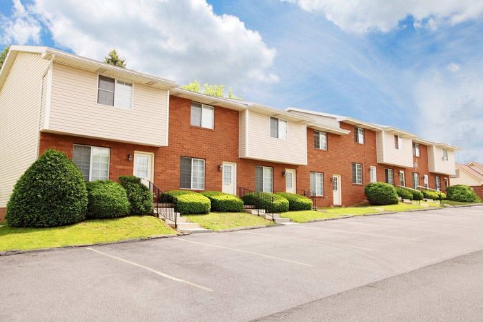 Altoona, PA, Apartments - Walton Heights - Property Exterior with Brick and Tan Walls, Concrete Steps with Railings, Manicured Grass Lawn and Bushes, and Parking Lot.