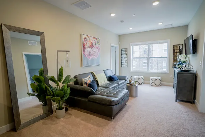 Apartments Near Penn State Harrisburg in Middletown, PA - Woodland Hills - Living Room with Carpeted Flooring, Black Sectional Couch, Wall Art, Coffee Table, Floor Lamp, Standing Mirror, Potted Plant, Storage Cabinet, Wall-Mounted TV, Ottoman Stools, and Large Window.
