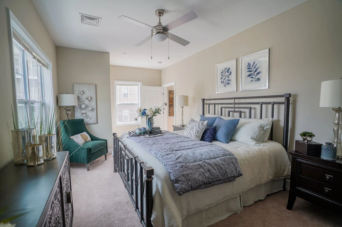 Pet-Friendly Apartments in Middletown, PA - Woodland Hills - Large Bedroom with Carpeted Flooring, Queen Sized Bed, Nightstands with Lamps, Dresser, Ceiling Fan, Armchair, Floor Lamp, and Windows.