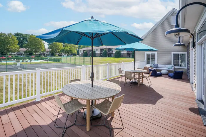 Pet-Friendly Apartments in Millersville PA - Millers Crossing - Outdoor Sundeck with Wooden Tables and Chairs with Blue Umbrellas, Outdoor Sectional Couch, Beanbags, Coffee Table, White Railing, and View of Tennis Court and Swimming Pool.