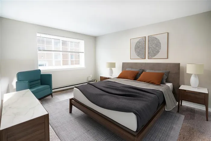 One-Bedroom Apartments in Norristown, PA - The Communities of Norris Hills - Furnished Bedroom with Carpeted Flooring, Area Rug, Large Bed, Marble-Topped Wooden Nightstands with Lamps, Wooden Dresser, Blue Armchair, White Walls, and Large Window.
