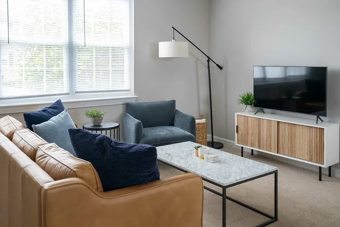 Wilmington, DE, Apartments - Greenview at Chestnut Run - Furnished Living Room with Carpeted Flooring, Leather Couch, Blue-Grey Armchair, Marble-Topped Coffee Table, TV Monitor with Wood and White Details, TV, Standing Lamp, Basket, Round Side Table, and Large Window.