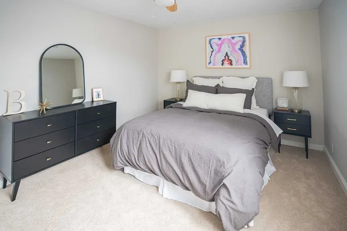 1-Bedroom Apartments in Wilmington, DE - Greenview at Chestnut Run - Spacious Bedroom with Carpeted Flooring, Large Bed, Dark Nightstands with Drawers and Lamps, Black Dresser, Framed Mirror, Ceiling Fan, and White Walls.