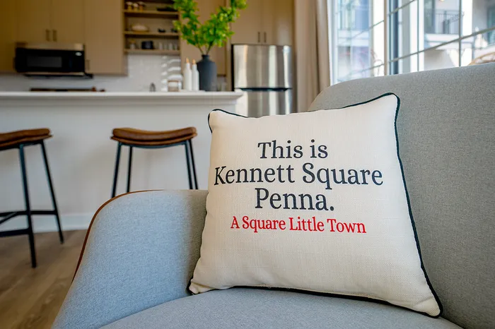 Bright living space featuring a gray armchair with a pillow that reads 'This is Kennett Square Penna,' with a view of the kitchen and barstools in the background.