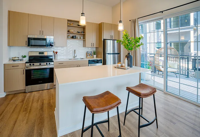 Modern kitchen with light wood cabinetry, stainless steel appliances, a white countertop island with barstools, and sliding glass doors leading to a patio.