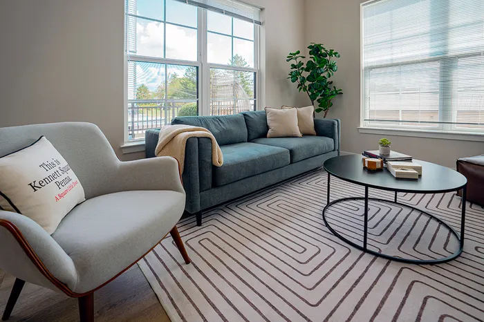 Spacious living room with a large gray sofa, a white armchair, a round coffee table, and large windows letting in natural light.