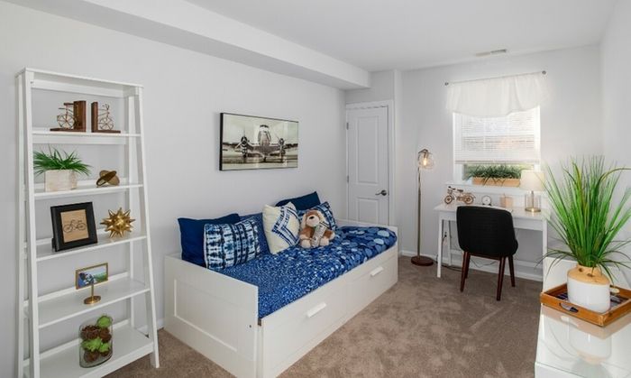 One-Bedroom Apartments in Bensalem, PA - Village Square - Bedroom with Carpeted Flooring, Bed, Wooden Dresser with Mirror, Green Armchair, Floor Lamp, and Large Window.