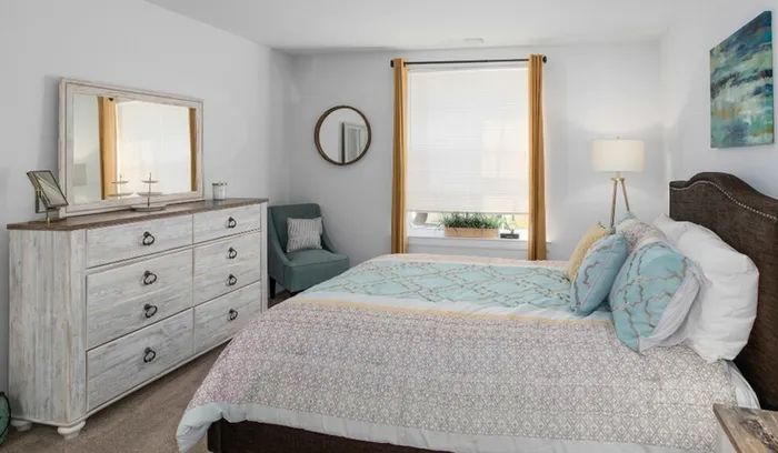 One-Bedroom Apartments in Bensalem, PA - Village Square - Bedroom with Carpeted Flooring, Bed, Wooden Dresser with Mirror, Green Armchair, Floor Lamp, and Large Window.