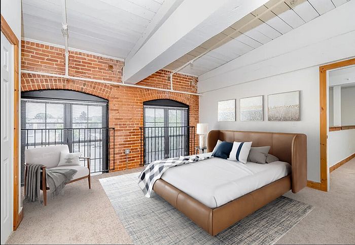 Apartments Near Da Vinci Science Center - The Mill - A Carpeted Bedroom with Brick Accent Walls, a Queen Sized Bed, and a Mirror-Dresser Set