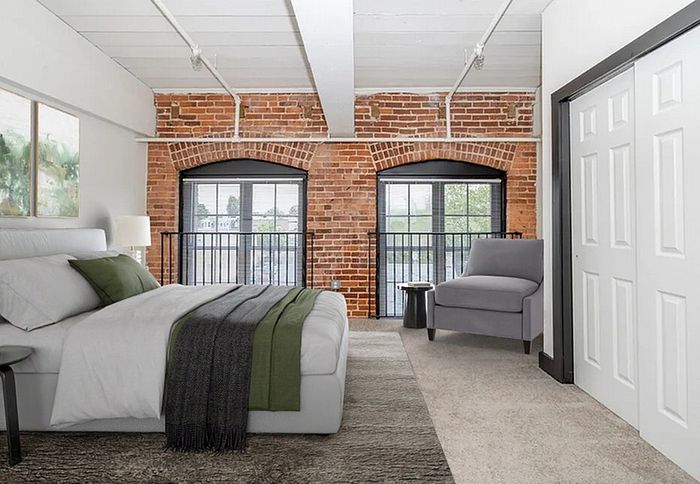 Apartments Near Da Vinci Science Center - The Mill - A Carpeted Bedroom with Brick Accent Walls, a Queen Sized Bed, and a Mirror-Dresser Set