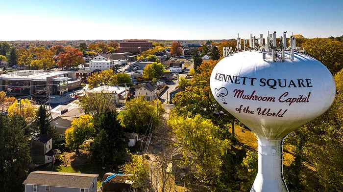 Panoramic aerial view of Kennett Square with the iconic water tower labeled 'Mushroom Capital of the World' surrounded by vibrant fall foliage.