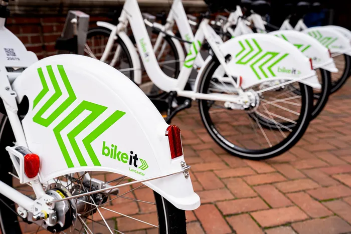 Row of modern white rental bikes with green branding, parked on a brick pathway.