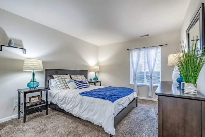 Inviting modern bedroom with plush carpet, a stylish bed with white and blue accents, and natural light through sheer curtains.