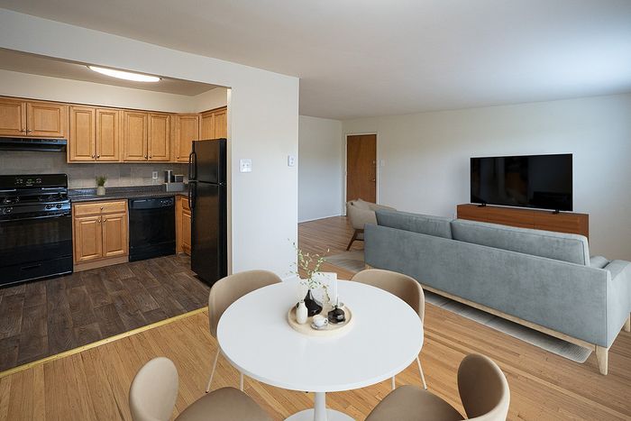 Modern open-concept apartment showcasing a cozy living room with a sleek grey sofa and TV, adjacent to a stylish kitchen with warm wooden cabinets and black appliances, complemented by a chic dining area with a round white table.