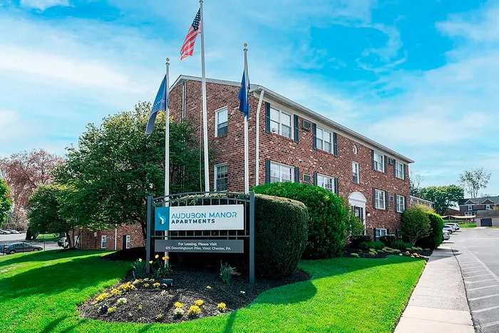 Charming Audubon Manor Apartments with classic brick architecture, lush landscaping, and a welcoming entrance under a clear blue sky.