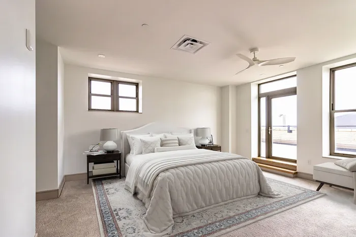 Bright and spacious bedroom with elegant white bedding, modern ceiling fan, and large windows offering natural light and balcony access.