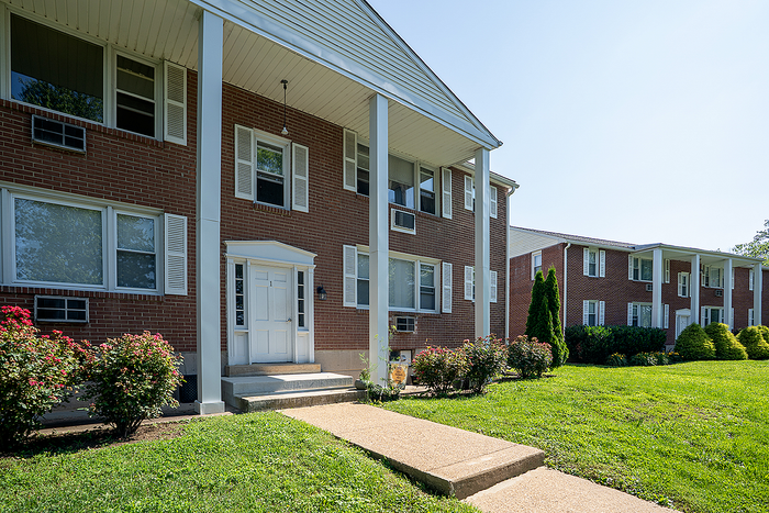 Charming brick apartment building with lush green landscaping, featuring classic white shutters and columns for a welcoming ambiance.