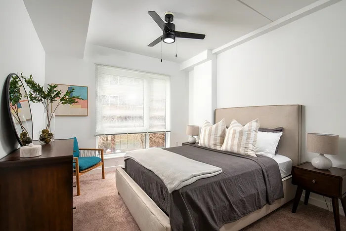 Inviting bedroom with elegant decor, featuring a plush bed, modern ceiling fan, cozy armchair, and abundant natural light through large windows.