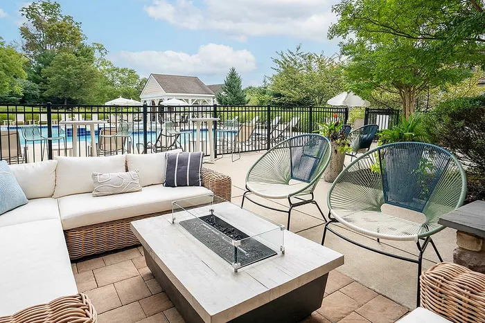 Modern outdoor lounge area with stylish wicker furniture, cozy fire pit, and a view of a sparkling pool surrounded by lush greenery.
