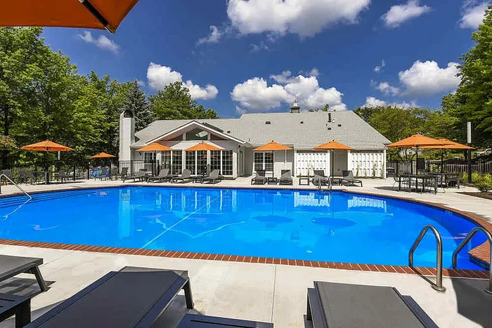 Inviting outdoor pool area with luxurious sun loungers, surrounded by lush greenery and a charming clubhouse under a bright blue sky.