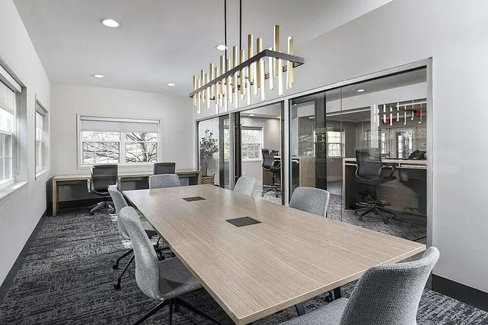 Elegant conference room with modern lighting, a spacious wooden table, ergonomic chairs, and floor-to-ceiling windows providing ample natural light.