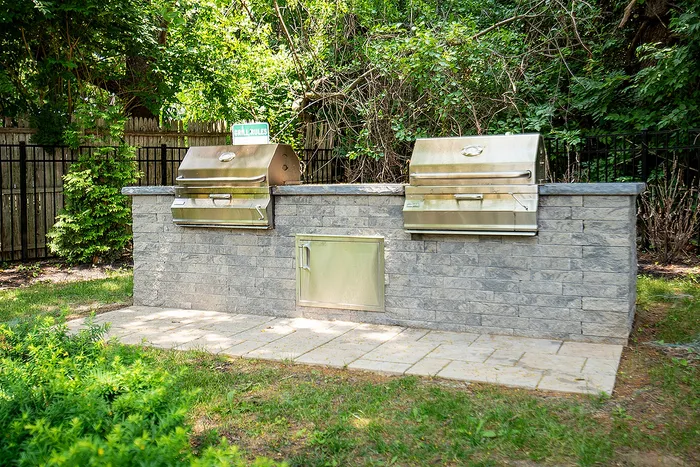 Inviting outdoor BBQ area with twin stainless steel grills set against a stylish stone backdrop, surrounded by lush greenery.